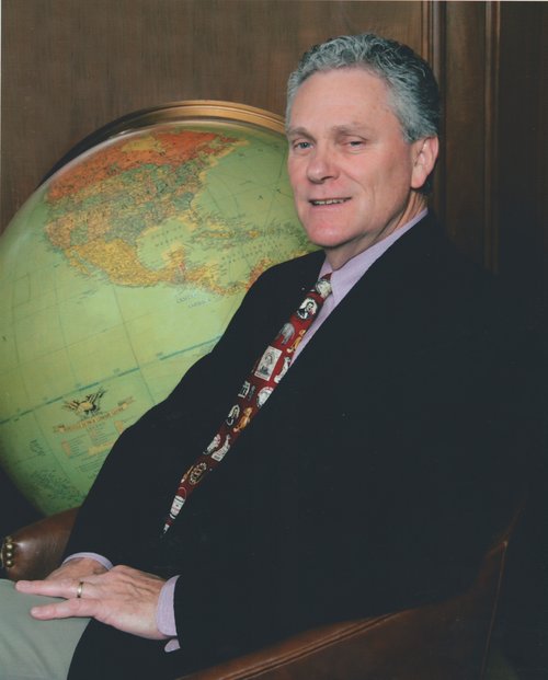 Martin Mack Teasley sits in a brown leather armchair in front of a large rotating globe. Teasley wears a black suit jacket, grey slacks, red tie with pictures of presidents on it, and a grey shirt.