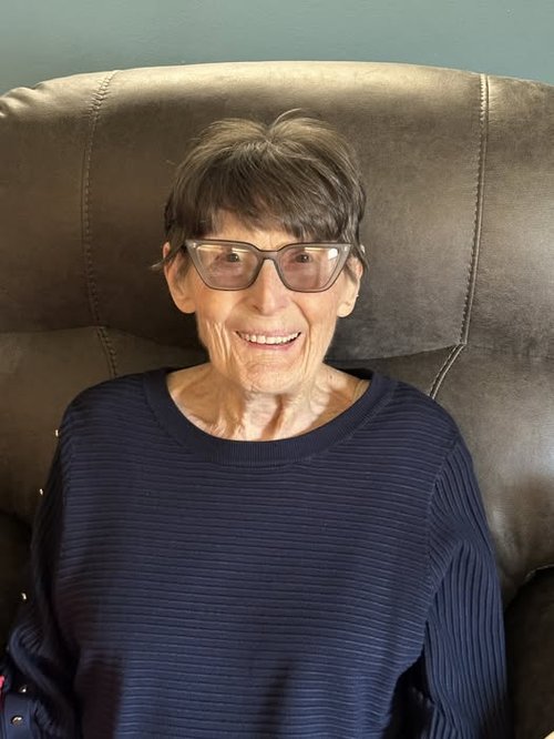 Joretta Duncan sits on a brown leatherette arm chair. She wears a purple sweater with horizontal stripes, brown cat-eye glasses and a smile.