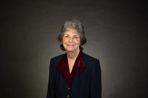 Marilee McBoyle-Wiesner stands in front of a lightly lit charcoal background. She wears a black jacket with maroon velvet collar folded out and gold buttons. She is smiling.