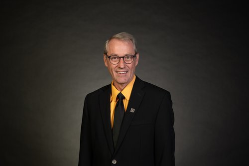 Sean Bird stands in front of a lit charcoal background wearing a black suit jacket, gold shirt, and black tie. He wears black, circular glasses and a slight smile.