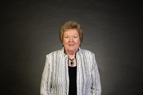 Shirley Antes stands in front of a lit charcoal background wearing a black and white patterned jacket, black shirt, and silver and black pendant necklace. Shirley wears black drop earrings and a slight smile.