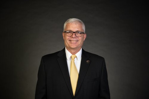 Steve Harmon stands in front of a lightly lit charcoal background. He wears a black suit jacket, white shirt, and yellow-gold tie. He has square black glasses and a smile.