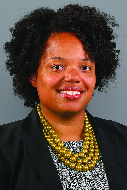 Natasha Oakes stands in front of a grey background smiling. Oakes wears a black blazer, grey patterned shirt, and gold necklace.