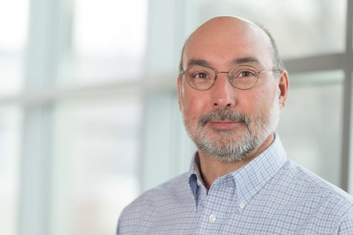 Dr. Tony Schountz stands in front of windows wearing a light blue plaid checkered shirt and grey circular glasses.