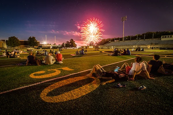 People sitting and reclining on the field of Welch Stadium watch fireworks burst in the air.
