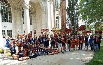 group of students hold up stringed instruments in front of stone building