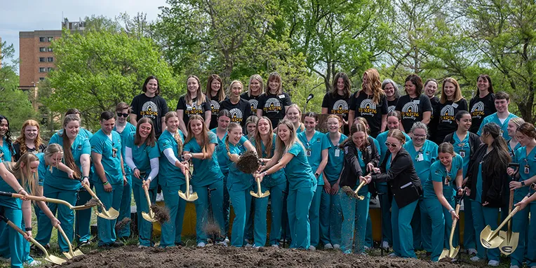 A group of college students and faculty members form a semicircle around a pile of dirt while holding shovels and scooping dirt.