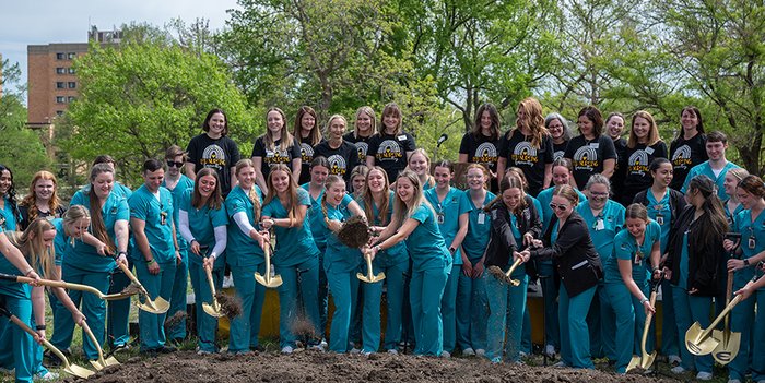 A group of college students and faculty members form a semicircle around a pile of dirt while holding shovels and scooping dirt.