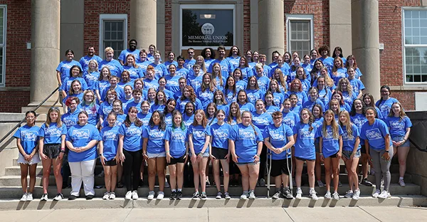 More than 90 students in blue T-shirts stand on steps of a building for a group photo.