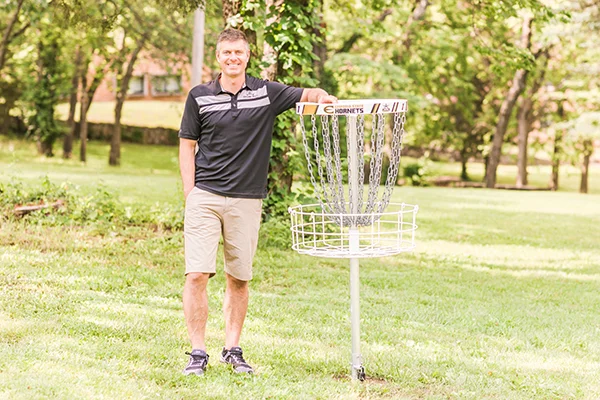 Dynamic Discs owner Jeremy Rusco posing in front of disc golf basket at ESU's Disc golf course, Hornet Hills