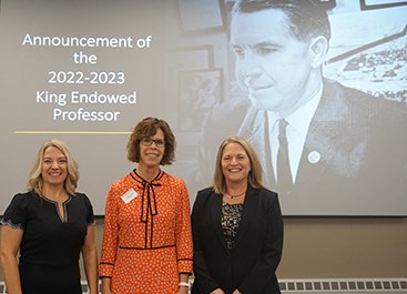 Three women stand in front of an image of an older man.